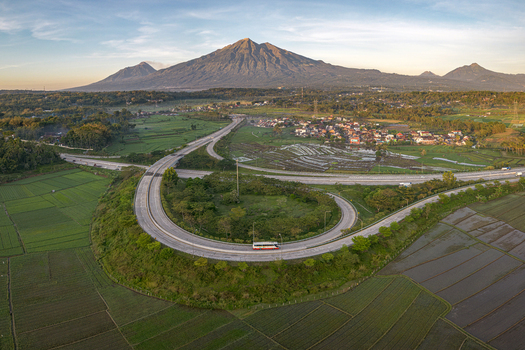 Aerial view of Jalan Tol Bawen Salatiga highway curve winding through green rice fields with a majestic mountain peak in the background Kecamatan Tengaran Jawa Tengah Indonesia.