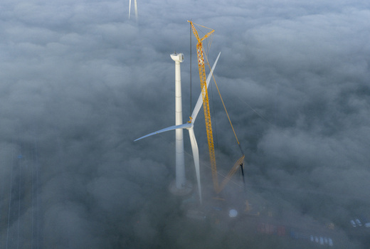 Aerial view of a white wind turbine and a yellow lattice boom crane emerging from dense morning fog.