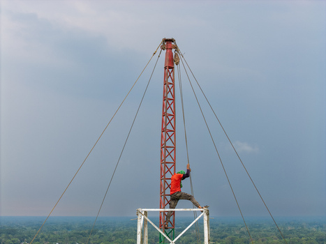 Trishal Upazila Bangladesh 13 May 2026 Aerial view of a worker climbing a tall red lattice mast with guy wires against a vast green landscape and cloudy sky. Trishal Upazila Bangladesh 13 May 2026 Aerial view of a worker climbing a tall red lattice mast with guy wires against a vast green landscape and cloudy sky.