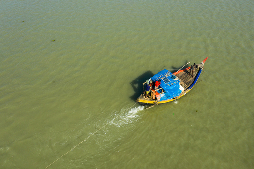 Aerial view of a traditional wooden fishing boat with a blue tarp cover and yellow hull carrying fishermen across the calm waters in Kuakata Barisal Division Bangladesh. Aerial view of a traditional wooden fishing boat with a blue tarp cover and yellow hull carrying fishermen across the calm waters in Kuakata Barisal Division Bangladesh.