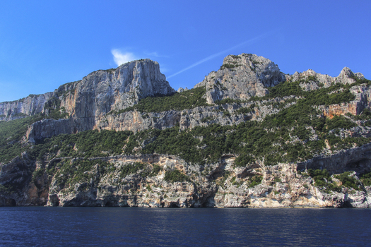View of the rugged limestone cliffs of the Gulf of Orosei rising above the deep blue sea under a clear sky in Italy. View of the rugged limestone cliffs of the Gulf of Orosei rising above the deep blue sea under a clear sky in Italy.