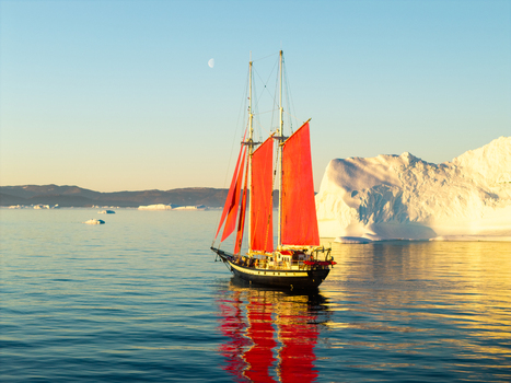 Aerial view of a sailboat with vibrant red sails navigating through calm waters surrounded by massive icebergs under a clear sky in Greenland. Aerial view of a sailboat with vibrant red sails navigating through calm waters surrounded by massive icebergs under a clear sky in Greenland.