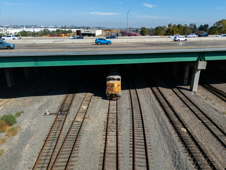 Aerial view of a freight train locomotive moving along tracks beneath a highway overpass with cars driving above under a clear blue sky in Carson California United States.