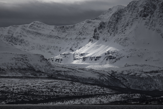 View of snow covered mountain peaks under a dramatic cloudy sky with a beam of sunlight hitting the slopes Tromso Troms Norway.
