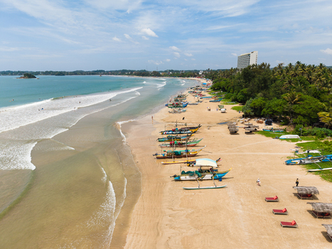 Aerial view of traditional fishing boats on a sandy beach with palm trees and a hotel under a blue sky in Weligama Southern Province Sri Lanka.