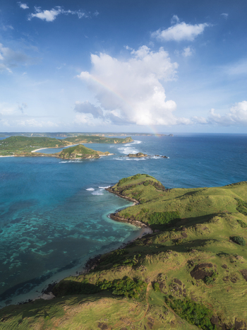 Aerial view of Tanjung Aan Beach with green hills turquoise ocean water and a rainbow under a blue sky in Kecamatan Pujut Nusa Tenggara Barat Indonesia.