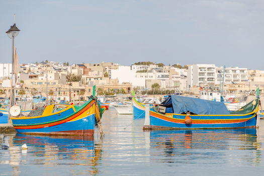 View of vibrant blue and yellow traditional fishing boats bobbing gently in the calm harbor waters against the backdrop of the white buildings Marsaxlokk Malta. View of vibrant blue and yellow traditional fishing boats bobbing gently in the calm harbor waters against the backdrop of the white buildings Marsaxlokk Malta.