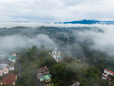 Aerial view of city buildings and houses on lush green hills partially covered by thick morning fog under a cloudy sky Kandy Central Province Sri Lanka. Aerial view of city buildings and houses on lush green hills partially covered by thick morning fog under a cloudy sky Kandy Central Province Sri Lanka.