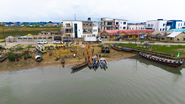 Aerial view of wooden boats moored along the muddy bank with people walking near the shore and modern buildings in the background in Minima Rivers Nigeria.
