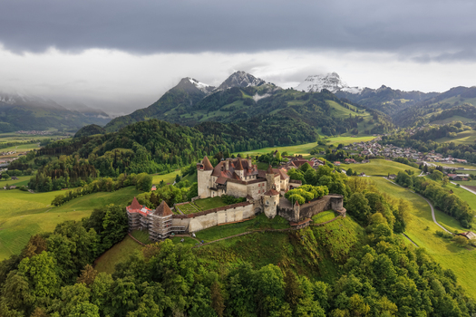 Aerial view of the historic Gruyeres Castle perched on a hilltop surrounded by lush green meadows dense forests and snow capped peaks in Gruyeres Fribourg Switzerland.