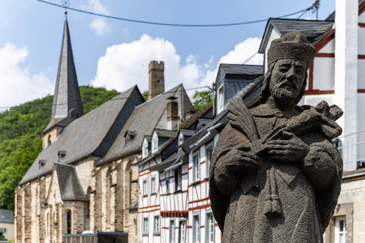 View of the stone statue of Saint John of Nepomuk with the historic Dreifaltigkeitskirche church and traditional half timbered houses under a blue sky in Monreal Rhineland Palatinate Germany. View of the stone statue of Saint John of Nepomuk with the historic Dreifaltigkeitskirche church and traditional half timbered houses under a blue sky in Monreal Rhineland Palatinate Germany.