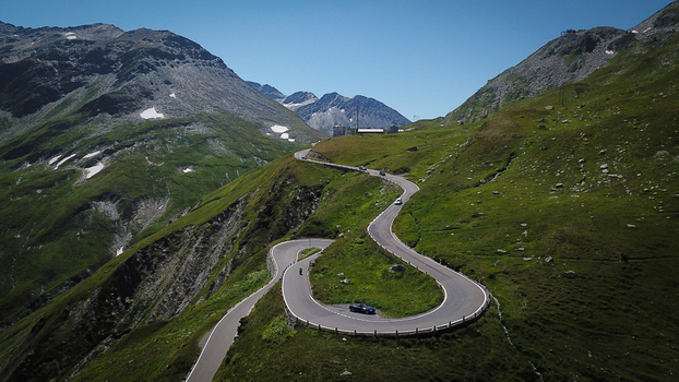 Aerial view of Furka Pass with winding roads and cars on green mountain slopes under a clear blue sky in Obergoms Valais Switzerland. Aerial view of Furka Pass with winding roads and cars on green mountain slopes under a clear blue sky in Obergoms Valais Switzerland.