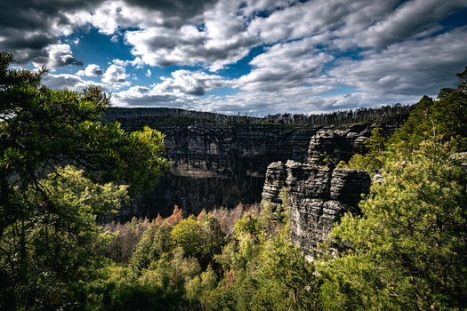 View of Pravcicka Brana sandstone arch formation surrounded by lush pine forests and dramatic rocky cliffs under a cloudy blue sky in Hrensko Usti nad Labem Region Czechia.