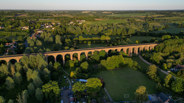 Aerial view of the historic Chappel Viaduct spanning the lush green Colne Valley with brick arches casting long shadows in the golden hour light Chappel England United Kingdom.