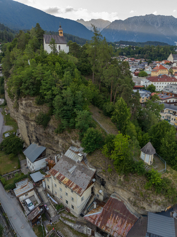 Aerial view of the historic church perched on a rocky cliff overlooking Imst with alpine mountain peaks and lush greenery surrounding the cliffside architecture in Imst Tyrol Austria.