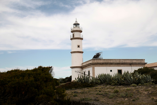 View of the historic lighthouse surrounded by agave plants and Mediterranean shrubs under a cloudy blue sky in Mallorca Balearic Islands Spain.