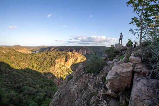 View of a woman standing on a rocky cliff edge looking over a vast sunlit gorge with lush green vegetation and distant hills in Kruger National Park South Africa.