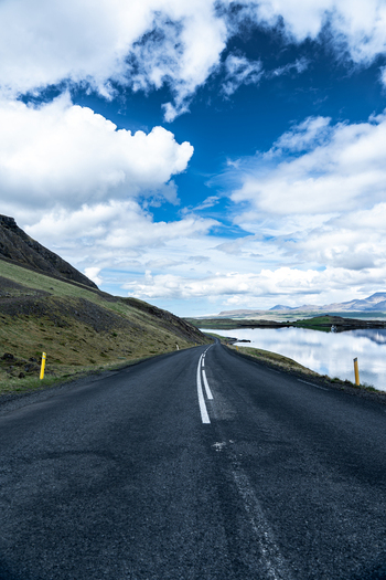 View of a winding asphalt road along the Hvalfjordur fjord with calm water reflections and grassy hills under a cloudy blue sky Iceland. View of a winding asphalt road along the Hvalfjordur fjord with calm water reflections and grassy hills under a cloudy blue sky Iceland.