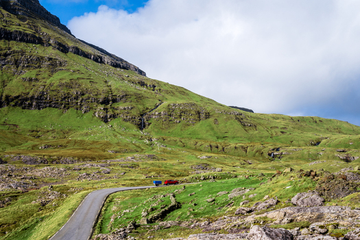 View of a red car with a trailer driving on a winding road through the mountainous green landscape of the Faroe Islands. View of a red car with a trailer driving on a winding road through the mountainous green landscape of the Faroe Islands.