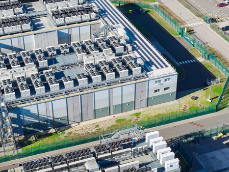Aerial view of a data center facility with rows of industrial cooling units on the roof surrounded by security fencing and paved access roads United Kingdom.
