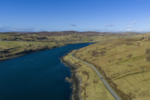 Aerial view of Gesto Bay with a winding coastal road and rolling hills under a clear blue sky in Isle of Skye Scotland United Kingdom.