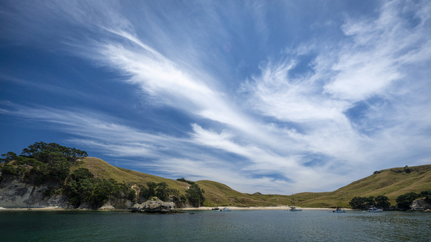 View of Coralie Bay with boats anchored in the calm water against rolling grassy hills and a bright blue sky with wispy clouds on Great Mercury Island Waikato Region New Zealand.
