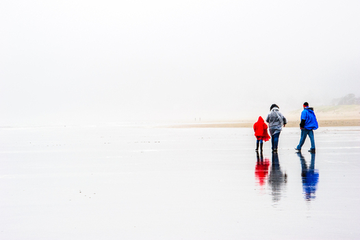 View of a child in a red raincoat and two adults walking along the wet reflective sand at Cannon Beach Oregon United States. View of a child in a red raincoat and two adults walking along the wet reflective sand at Cannon Beach Oregon United States.