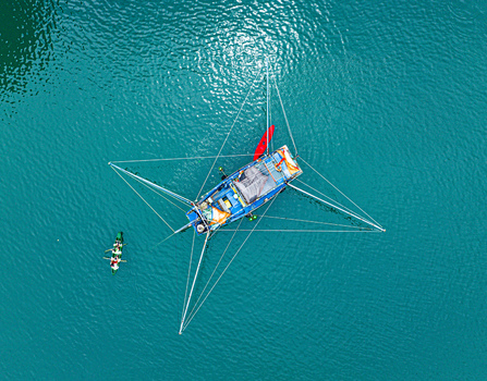 Aerial view of a traditional fishing boat with extended outriggers and a small kayak nearby floating on the turquoise waters of Ha Long Bay Vietnam.