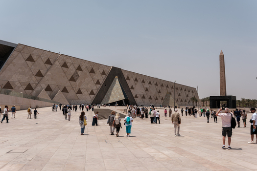 Giza Egypt 16 May 2026 View of the Grand Egyptian Museum with its triangular patterned facade and an ancient obelisk under a clear sky with tourists walking on the plaza.