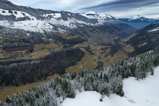 Aerial view of the Les Rives area with snow covered mountain slopes dense pine forests and traditional alpine chalets under a cloudy winter sky in Champery Valais Switzerland.