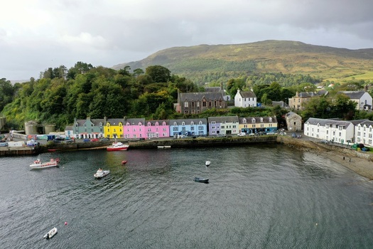 Aerial view of Portree Harbour featuring colorful houses along the waterfront with boats moored in the bay and rolling green hills in the background in Portree Scotland United Kingdom.