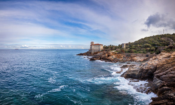 View of Castello del Boccale perched on a rocky cliff overlooking the blue Mediterranean sea with crashing waves and green hills in Calafuria Toscana Italy.