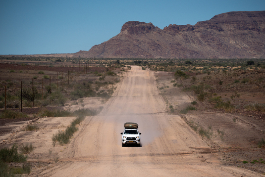 View of a white vehicle kicking up dust on a long straight dirt road towards distant mountains under a clear blue sky Keetmanshoop Karas Region Namibia. View of a white vehicle kicking up dust on a long straight dirt road towards distant mountains under a clear blue sky Keetmanshoop Karas Region Namibia.