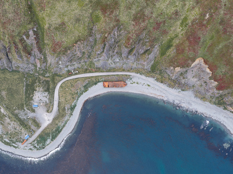 Aerial view of a rusted shipwreck resting on a gravel beach beside a winding coastal road and steep cliffs in Unalaska Alaska United States.