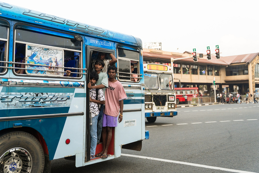 Sri Lanka 15 January 2026 View of young men hanging out of the open door of a crowded blue bus on a busy street near a terminal.