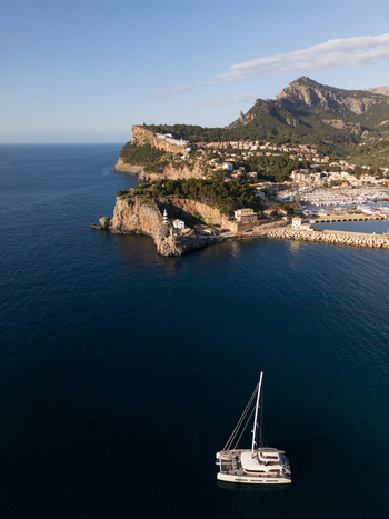 Aerial view of Port de Soller with a lighthouse on a rocky cliff and a white catamaran sailing on the deep blue sea Port de Soller Balearic Islands Spain.