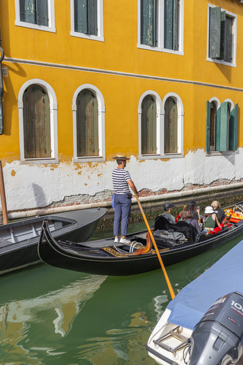Venice Italy 21 September 2026 View of a gondolier in a striped shirt and straw hat rowing a gondola with tourists along a canal past a yellow building. Venice Italy 21 September 2026 View of a gondolier in a striped shirt and straw hat rowing a gondola with tourists along a canal past a yellow building.