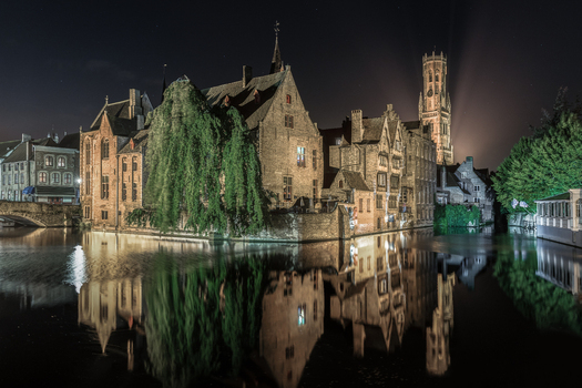View of historic medieval buildings and the illuminated Belfry tower reflected in the calm canal waters at night in Bruges Flanders Belgium.