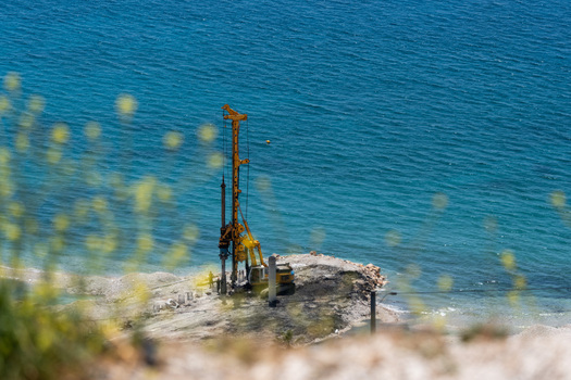 View of a yellow drilling rig operating on a rocky shoreline by the turquoise sea with blurred yellow flowers in the foreground Vlore County Albania. View of a yellow drilling rig operating on a rocky shoreline by the turquoise sea with blurred yellow flowers in the foreground Vlore County Albania.