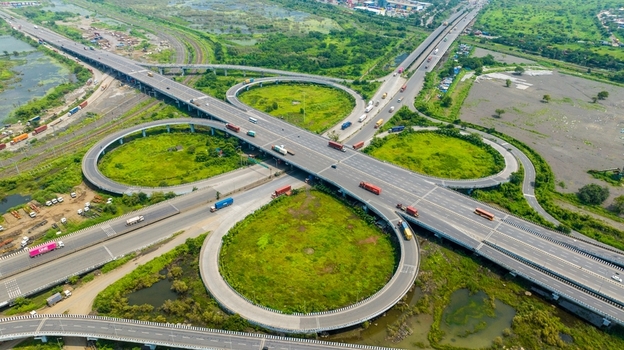 Aerial view of the JNPT Interchange with heavy traffic and trucks navigating the multi level highway surrounded by lush greenery and wetlands in Navi Mumbai Maharashtra India.