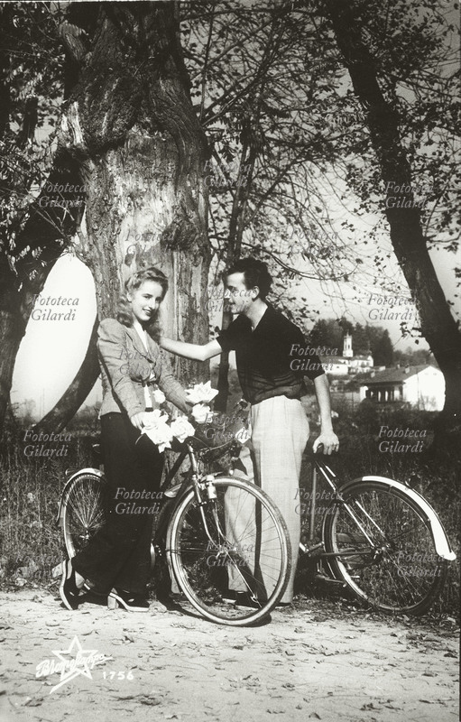 COPPIA di innamorati in gita in bicicletta lungo il fiume; sul mezzo della giovane donna appare una piccola bandierina italiana con lo stemma dei Savoia. Fotografia, Torino 1941