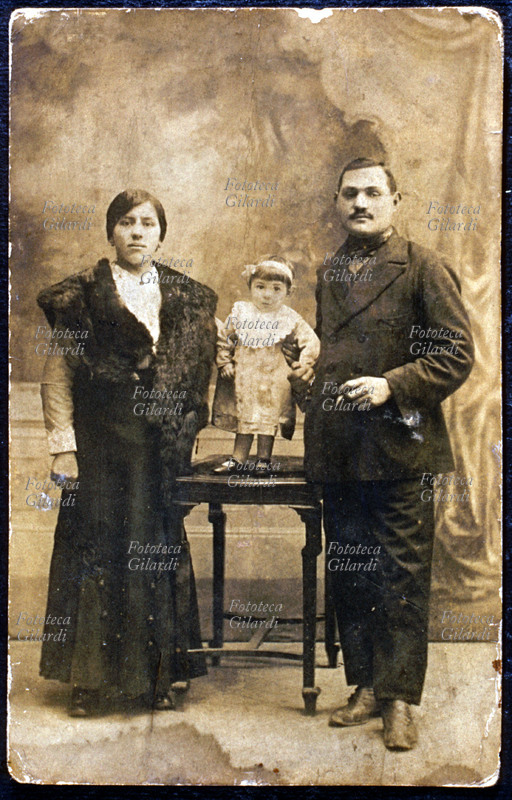 FAMIGLIA in posa: i due genitori in piedi, la minuscola ed elegante bambina tra loro sopra il tavolo. Fotografia di anonimo eseguita in atelier, Italia 1890 circa.