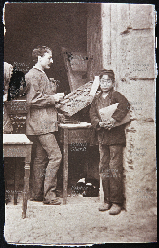 MESTIERI Tipografo e giovane apprendista davanti alla cassa di caratteri mobili; un cagnolino dorme acciambellato sotto il tavolo. Fotografia, Italia primi anni del 1900.