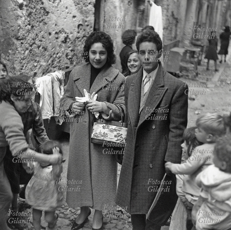 FIDANZATI giovani in posa nel vicolo, vestiti elegantemente, circondati da numerosi bambini. La coppia sorride ed ha un\