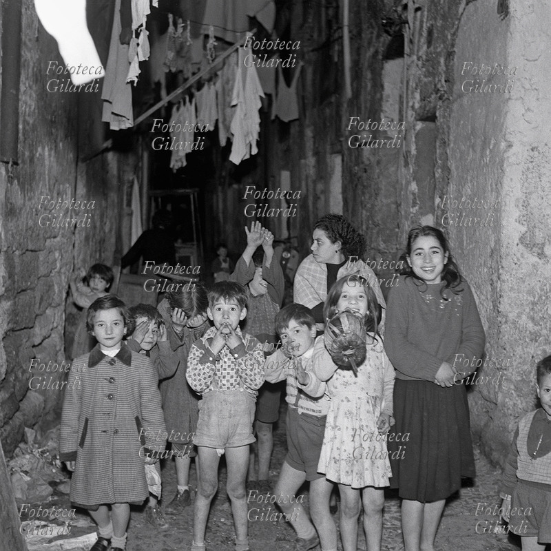 BAMBINI gruppo in posa, con una madre, a Cortile Cascino: nel buio del vicolo il lampo del flash provoca in loro diverse reazioni: dal piccolo spavento al divertimento, allo stupore; interessanti le varie espressioni. Fotografia di Ando Gilardi (1921 - 2012) #andogilardi da ampio servizio, Palermo 1957.