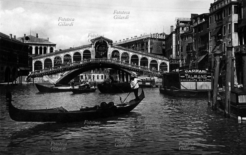 VENEZIA veduta del ponte di Rialto (realizzato da Antonio da Ponte nel 1591) con passaggio di gondole sul Canal grande; sulle chiatte chiuse attraccate alle fondamenta, estensione per collocare i tavolini dei bar, appaiono le scritte pubblicitarie "Bitter Campari" e "Talmone cioccolato al latte". Fotografia, Italia 1930 circa