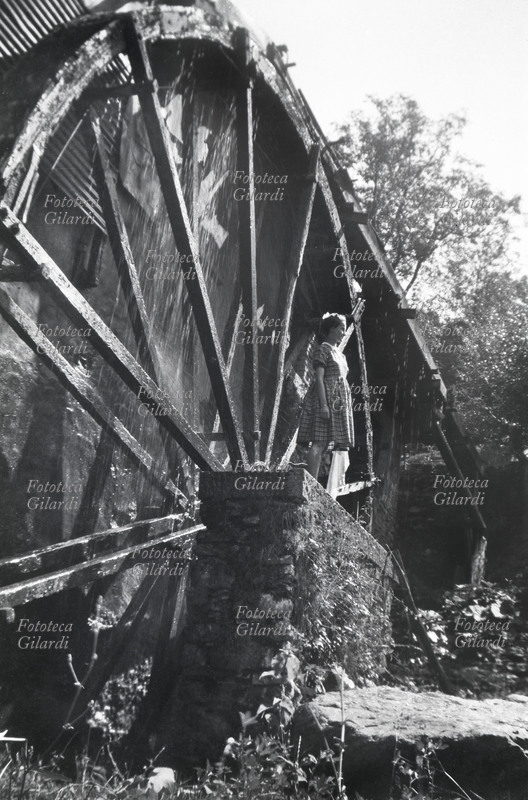 MULINO AD ACQUA la ruota in primo piano con accanto una ragazzina ritratta in piedi, per evidenziare la dimensione davvero enorme della macchina. L\