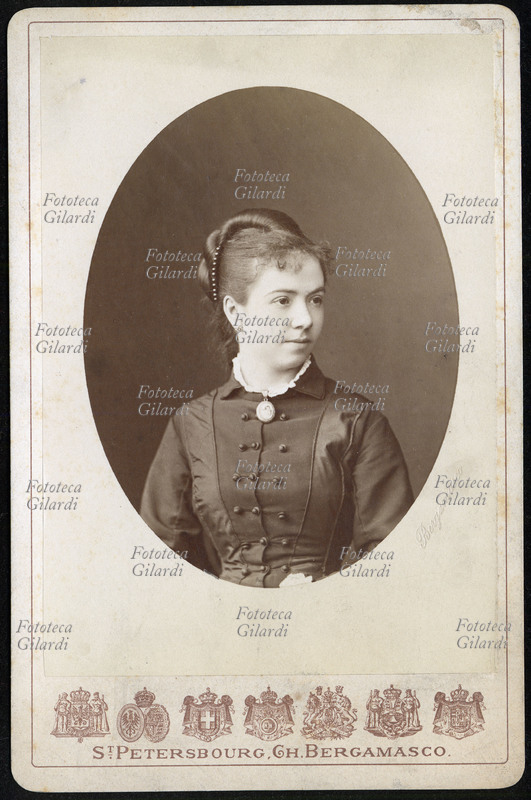 MODA Ovale con ritratto di giovane donna. Acconciatura femminile: coda ritorta che parte dal culmine della testa, appena sopra la fronte e che va a legarsi sulla nuca, arricchita da una collana di perle di colore scuro. Fotografia dello studio Charles Bergamasco (1830 - 1896), fotografo della corte imperiale; San Pietroburgo, Russia, 1878 circa.