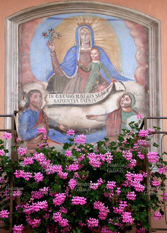 MADONNA AND CHILD in Glory and two Saints, honored by a lush cascade of pink ivy geraniums. The Child holds a scroll with the motto \
