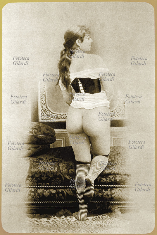 EROTISMO giovane donna, ripresa di schiena per evidenziare le natiche prosperose: indossa calze a righe e camicia bianca trattenuta da un bustino scuro. Francia 1885